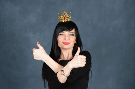 Expressive and glamorous beautiful girl, brunette in a black dress and the crown, showing his fingers up as a symbol of luck and success. Beautiful and slender woman looks at the camera, smiles, studio portrait on a monochrome blue, trendy shabby backgroundの写真素材