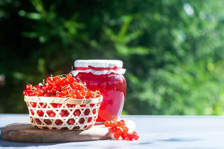 Red, juicy berries of red currants and jars of berry jam on a wooden table, on a green background of the garden, harvest and cooking theme, space for textの写真素材