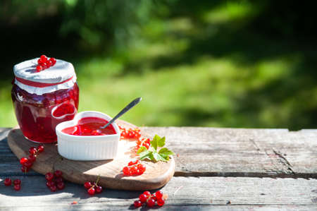Red, juicy berries of red currants and jars of berry jam on a wooden table, on a green background of the garden, harvest and cooking theme, space for textの写真素材