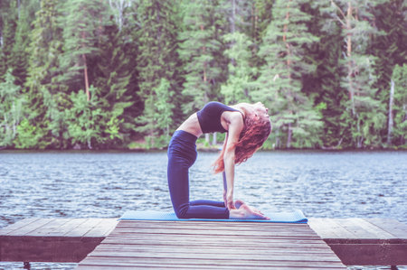 Young yogi girl practicing yoga, stretching in Ustrasana exercise, Camel pose, on the lake. Concept of healthy life and natural balanceの写真素材