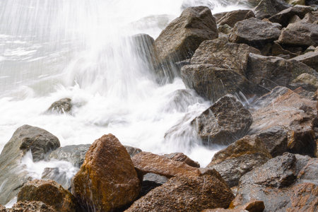 Powerful sea waves crash against coastal rocks, creating splashes and white foam. A dynamic seascape scene capturing the strength and movement of nature at the water's edge.の写真素材