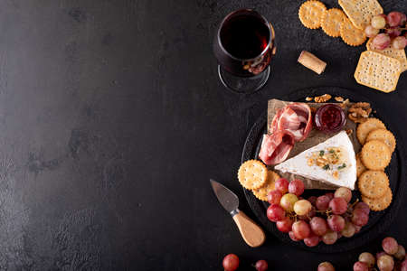 piece of brie cheese with grapes, crackers and red wine on a black tableの写真素材