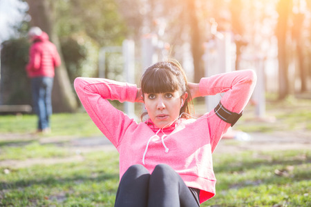 Young Woman Doing ABS Exercises at Park. She is also Listening Music with Earphones. The girl is sitting on a Fitness Mat. Some People on Background doing Exercises too.の写真素材