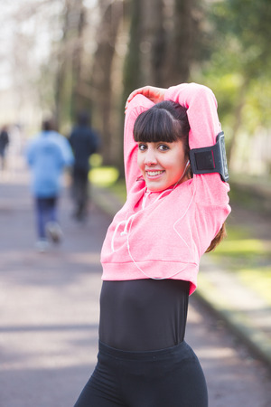 Young Woman Doing Arms and Shoulders Stretching Exercises. She Has a Smart Phone Holder on the Left Arm and she is Listening Music with Earphones. Urban Park setting.の写真素材