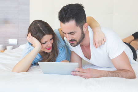 Young Couple with Digital Tablet on Bed. They are in a Modern Hotel Room and They wear Underwear. The Man is holding the Tablet with his left hand. Technology addiction and love concepts.の写真素材