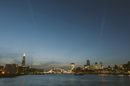 London skyline at dusk with River Thames on foreground. From left to right The Shard, Tower Bridge, Walkie Talkie and other skyscrapers. All buildings have lights on.の写真素材