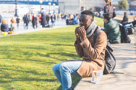 Young man having lunch break in London next to Tower Bridge. He is seated on a concrete low wall and...