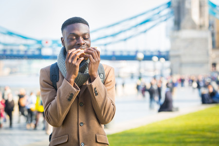 Young man having lunch break in London next to Tower Bridge. He is seated on a concrete low wall and...