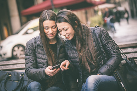 Two female twins looking at a smart phone. They wear black jackets and jeans. Urban scene with houses and street on background.の写真素材