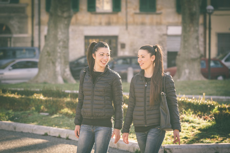Female twins walking at city public square. They wear black jackets and jeans. Buildings and cars on background. They walk side by side talking and looking around.の写真素材