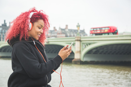 Beautiful redhair woman listening music in London with Westminster bridge on background. She wear...
