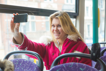 Beautiful young woman taking a selfie while commuting in London with double-decker bus. She is sitting next to the window with buildings and traffic on background.の写真素材