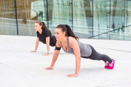 Two women doing push-ups exercises in a urban context. They are wearing gray and black sportswear