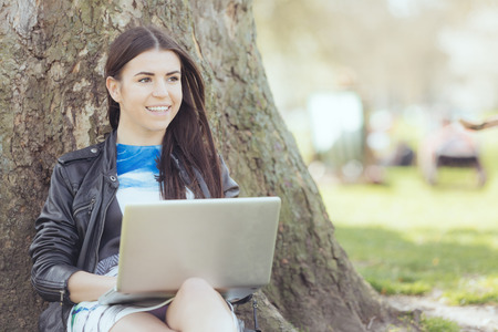 Young woman using computer at park in London. She is lying next to a tree, resting in the shadow on a London sunny day in spring.の写真素材
