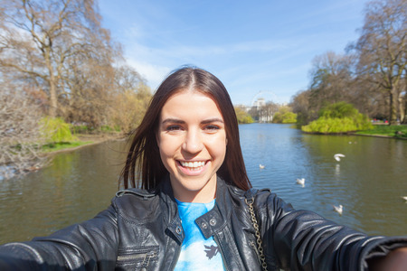 Beautiful young woman taking a selfie in London at St James park with lake and trees on background. の写真素材