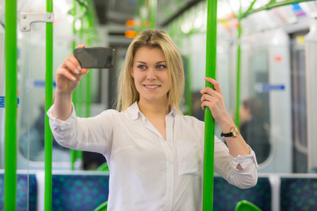Beautiful blonde young woman taking a selfie with smart phone in London tube train. She is holding to a pole, wearing a white shirt and looking at the phone.の写真素材