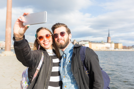 Young hipster couple taking a selfie in Stockholm with sea and old town on background.  They are both wearing sunglasses and looking at smart phone.の写真素材