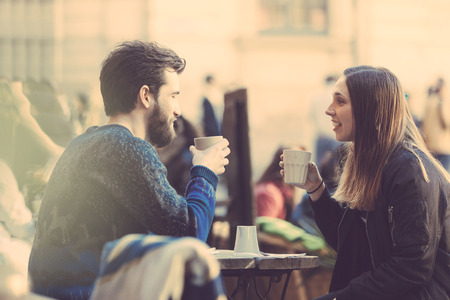 Hipster couple drinking coffee in Stockholm old town. They're sitting face to face. The man is wearing a blue sweater and the woman a striped shirt with black leather jacket. See-through shot.の写真素材