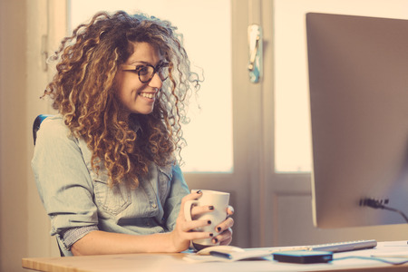 Young woman working at home or in a small office, vintage hipster clothing, curly hair. Cup of tea or coffee on the desk with some technological devices.の写真素材