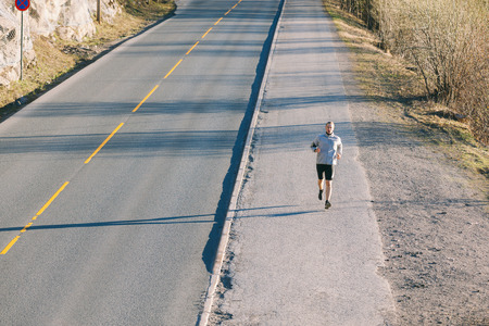 Young man running outside along an empty mountain road in the early morning. He has nordic features, and he's wearing black shorts and a gray sweater.の写真素材