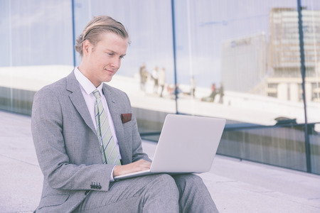 Fashioned young man working with laptop outdoor. He has nordic facial features, and wearing a light gray suit. There is a modern background with white stones and big windows.の写真素材