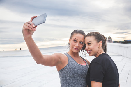 Two women taking a selfie after workout. They are wearing gray and black sportswear, modern urban background, cloudy day.の写真素材