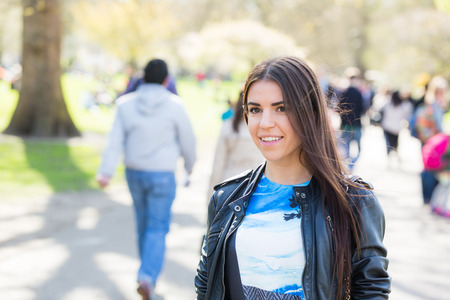 Young woman walking at park in London. She is wearing a short dress with a black jacket, there are some unrecognizable persons on backgroundの写真素材
