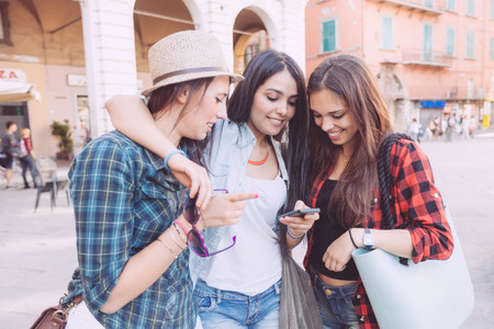 Three young women with smart phone in the city, talking each other and smiling. This is a mixed race group, one girl is half asian and one is middle eastern. Lifestyle, friendship and urban life concepts.の写真素材