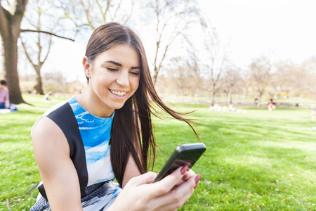 Young woman looking at smart phone at park in London. She is lying on the grass, resting on a London spring sunny day.の写真素材