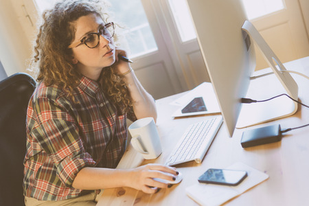 Young woman working at home or in a small office, vintage hipster clothing, curly hair. She seems to be stressed or thoughtful, There is a cup of tea or coffee on the desk with some technological devices.の写真素材