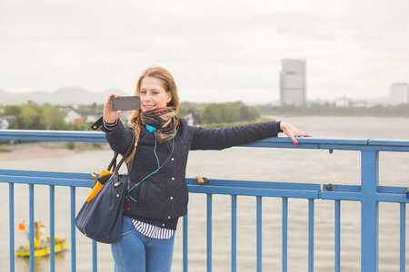 German girl taking selfie in Bonn with Rhein on background. She is standing next to the railing, holding the smart phone with one hand and looking at it.の写真素材