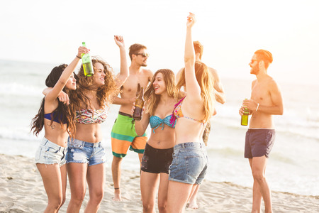 Multiracial group of friends having a party on the beach, dancing and drinking beer. They are teenagers, four girls and three boys, standing just next to the seaside.の写真素材
