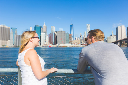 Caucasian couple visiting New York in summer. They are looking at sckyscrapers of lower Manhattan and showing their back to the camera. They have blond hair and wear light colored clothes.の写真素材