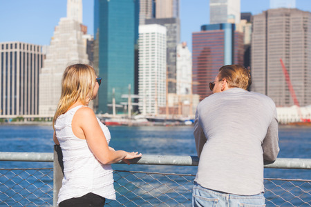 Caucasian couple visiting New York in summer. They are looking at sckyscrapers of lower Manhattan and showing their back to the camera. They have blond hair and wear light colored clothes.の写真素材