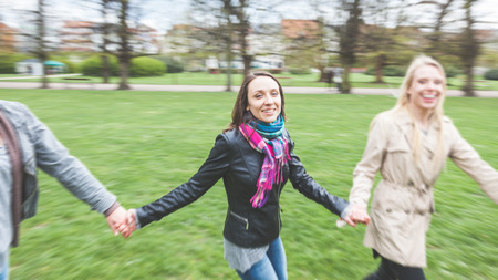 Group of friend at park in Copenhagen. Three women are holding their hands and running in circle together. They are very happy and smiling. There are grass, trees and some houses on the background.の写真素材