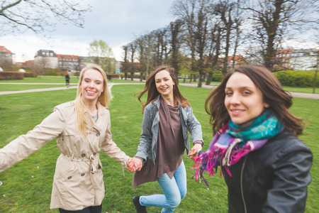 Group of friend at park in Copenhagen. Three women are holding their hands and running in circle together. They are very happy and smiling. There are grass, trees and some houses on the background.の写真素材
