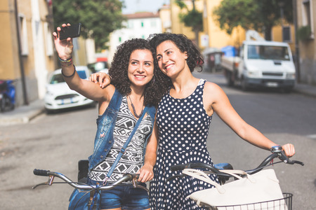 Female friends taking a selfie with their bicycles in the city. They are two women, a couple or sisters, wearing summer clothes and looking at their smart phone. There are some houses on background.の写真素材