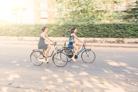 A couple of friends riding bikes on the street. Two women wearing summer clothes are riding bikes.  There are some trees on the background. selective blur with panning technique.の写真素材