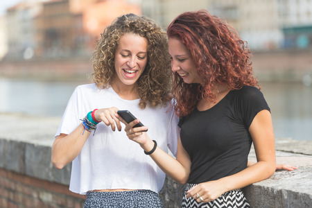 Two girls looking at smart phone. A couple of friends is standing next to the river. They are smiling and having fun together looking at their smartphone. There are houses on the background.の写真素材