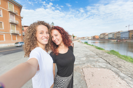 Two girls taking a selfie with Italian cityscape on background. They are standing next to the river and taking a self portrait while smiling and having fun together.の写真素材