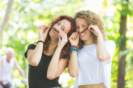 Two girls making moustache with their hair.  They are happy and wearing summer clothes, with some blurred green trees on background.の写真素材