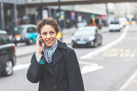 Young woman talking on the phone next to a busy street in London. She is a beautiful mixed race woman on her late twenties. Lifestyle and communications concepts in a urban context.の写真素材