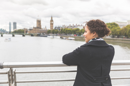 Young mixed race woman on a bridge in London. She is wearing a black coat and leaning on the railing while looking at Thames river and Big Ben on background. Tourism and lifestyle concepts.の写真素材