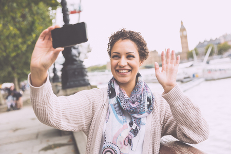 Beautiful mixed race woman taking a selfie in London with Thames river and Big Ben on background. She is holding the camera and waving with her hand.の写真素材
