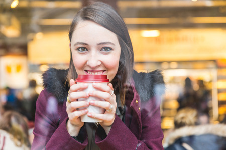 Beautiful young woman in a cafe holding a cuf of tea, seen through the window with buildings and lights reflections. She is looking at camera. Lifestyle and loneliness concepts.の写真素材