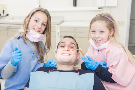 Two little dentist and dental assistant with a examining mouth of an adult patient. They are very young girls and the patient is a man. This is a funny  and surreal situation.の写真素材
