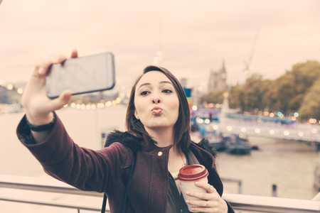 Beautiful young woman taking a selfie in London with Big Ben on background. She is holding the camera with one hand and looking at it smiling. She is also holding a cup of tea on the other hand.の写真素材