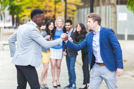 Two business men compete in arm wrestling with some colleagues inciting them. They are a black man...