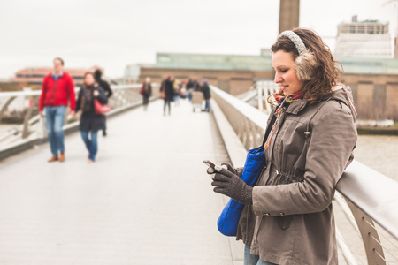 Beautiful woman typing on smart phone in London. She is leaning on the railing of a bridge, wearing warm clothes on a cold winter day. Travel, lifestyle and technology concepts.の写真素材