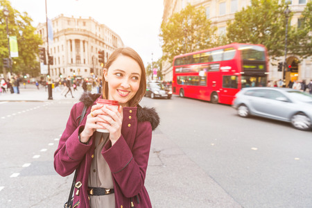 Woman in London standing by a busy road holding coffee. She is on her mid twenties and looking away from camera. Blurred traffic on background. Travel and lifestyle concepts.の写真素材